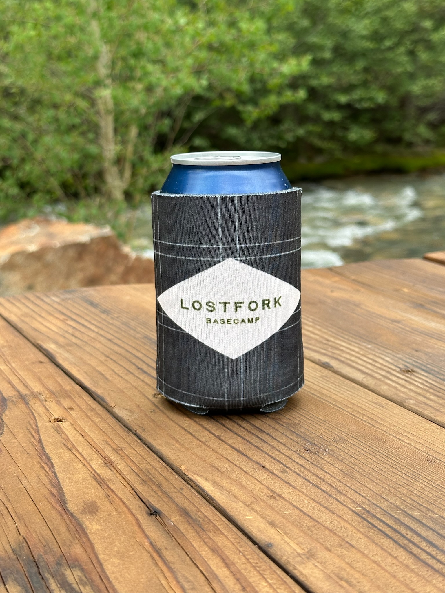 A blue can in a gray plaid koozie labeled "LOSTFORK BASECAMP" sits on a wooden table outdoors, with a stream and green foliage in the blurred background.