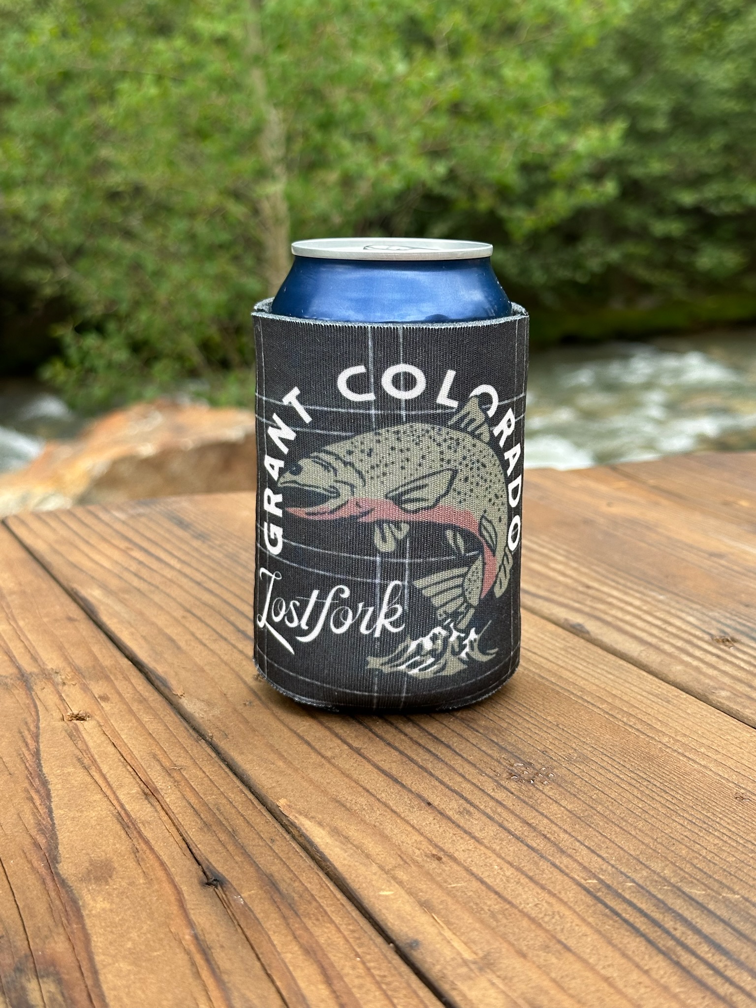 A blue can in a black koozie with a fish illustration and the text "Grant Colorado Lostfork" sits on a wooden surface outdoors near a river and greenery.