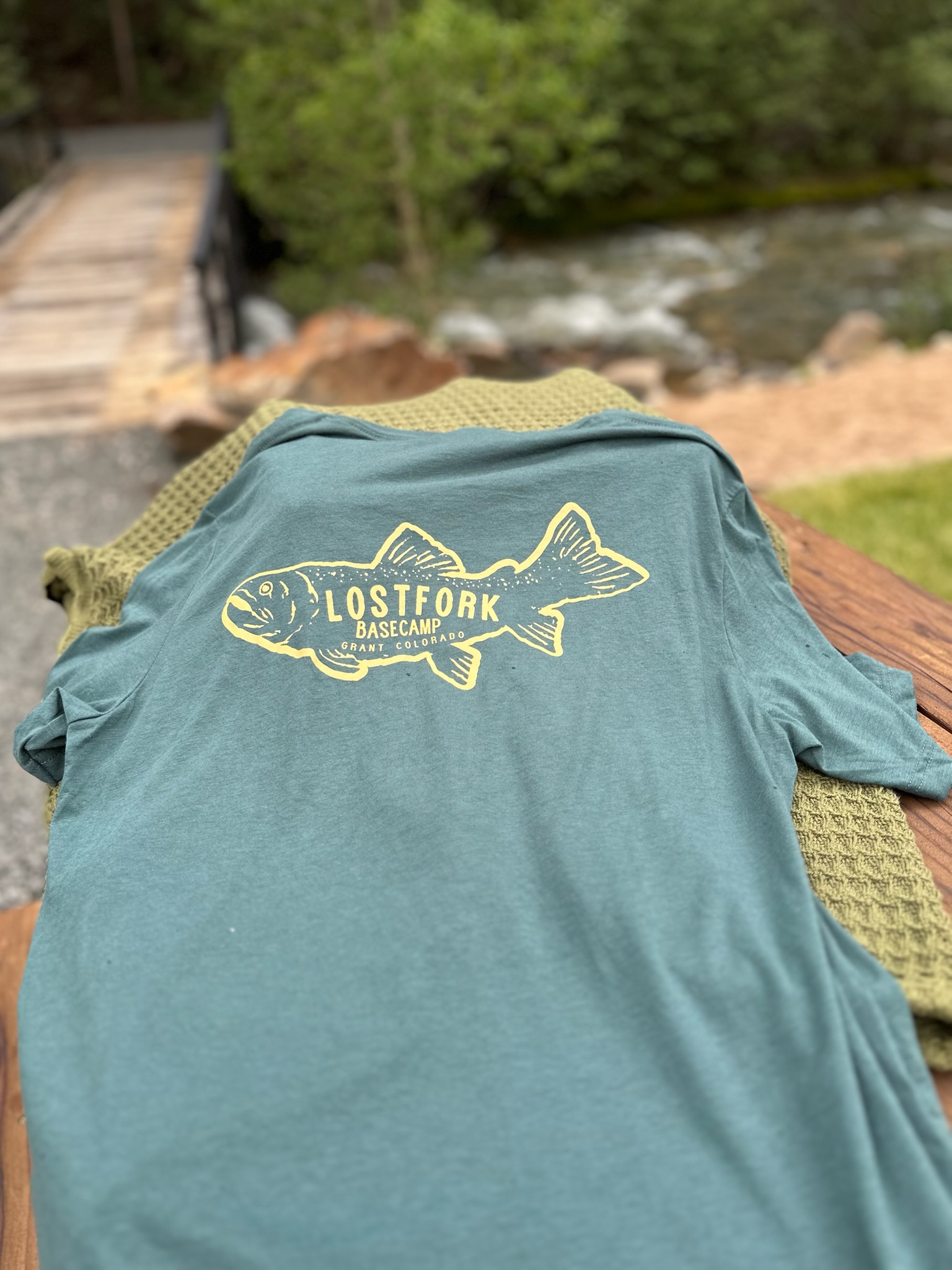 A green T-shirt with a yellow fish graphic and the words “Lost Fork Basecamp Stay Cozy” is laid out on a wooden surface outdoors, with a green textured blanket and a stream with trees in the background.