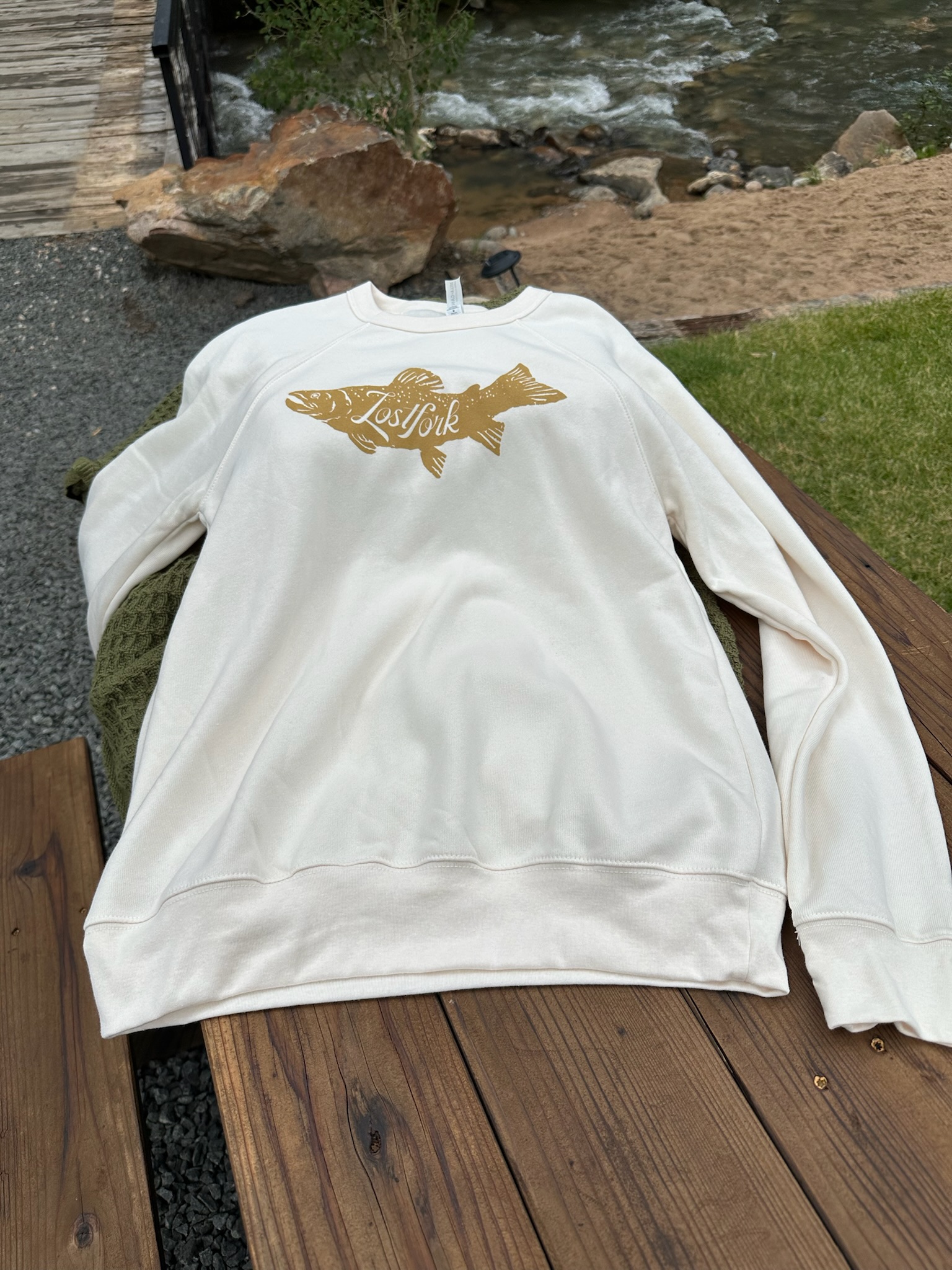 A white sweatshirt with a gold fish graphic and the word "Lofsjök" printed on it is laid out on a wooden bench near a river with rocks, sand, and greenery in the background.