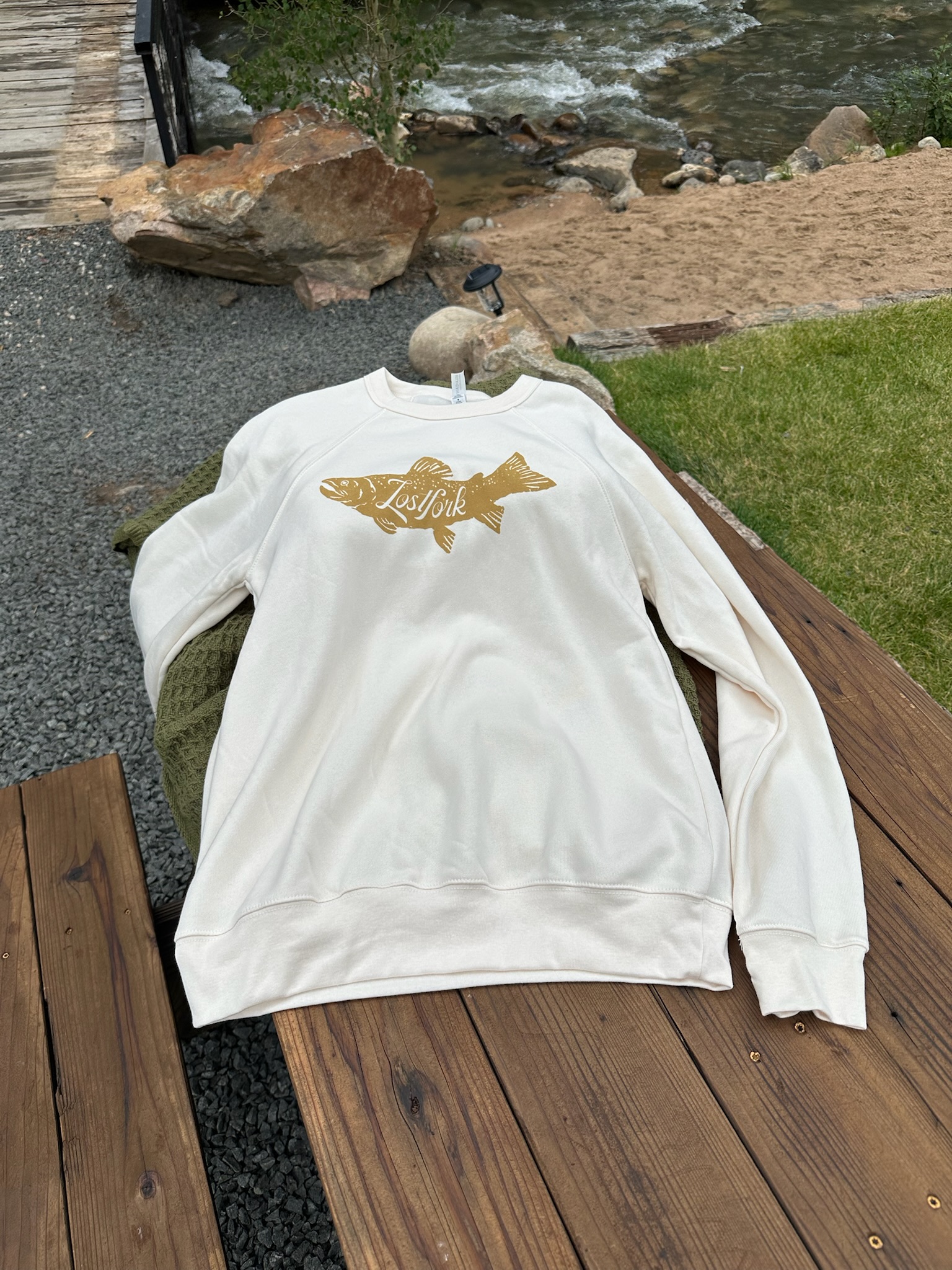 A white sweatshirt with a gold fish graphic and the words "Lily Lake" is laid out on a wooden bench outdoors near gravel, grass, rocks, and a flowing creek.