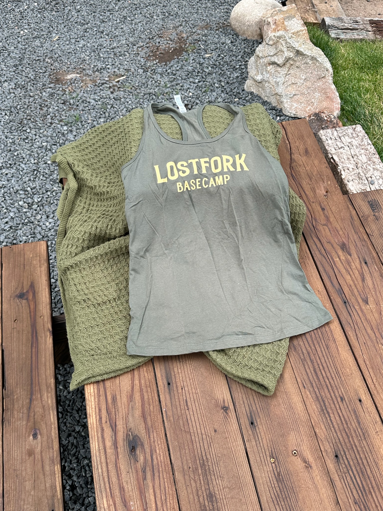 An olive green tank top with "LOSTFORK BASECAMP" in yellow letters is laid on top of a textured green blanket, both resting on a wooden bench with gravel and rocks in the background.
