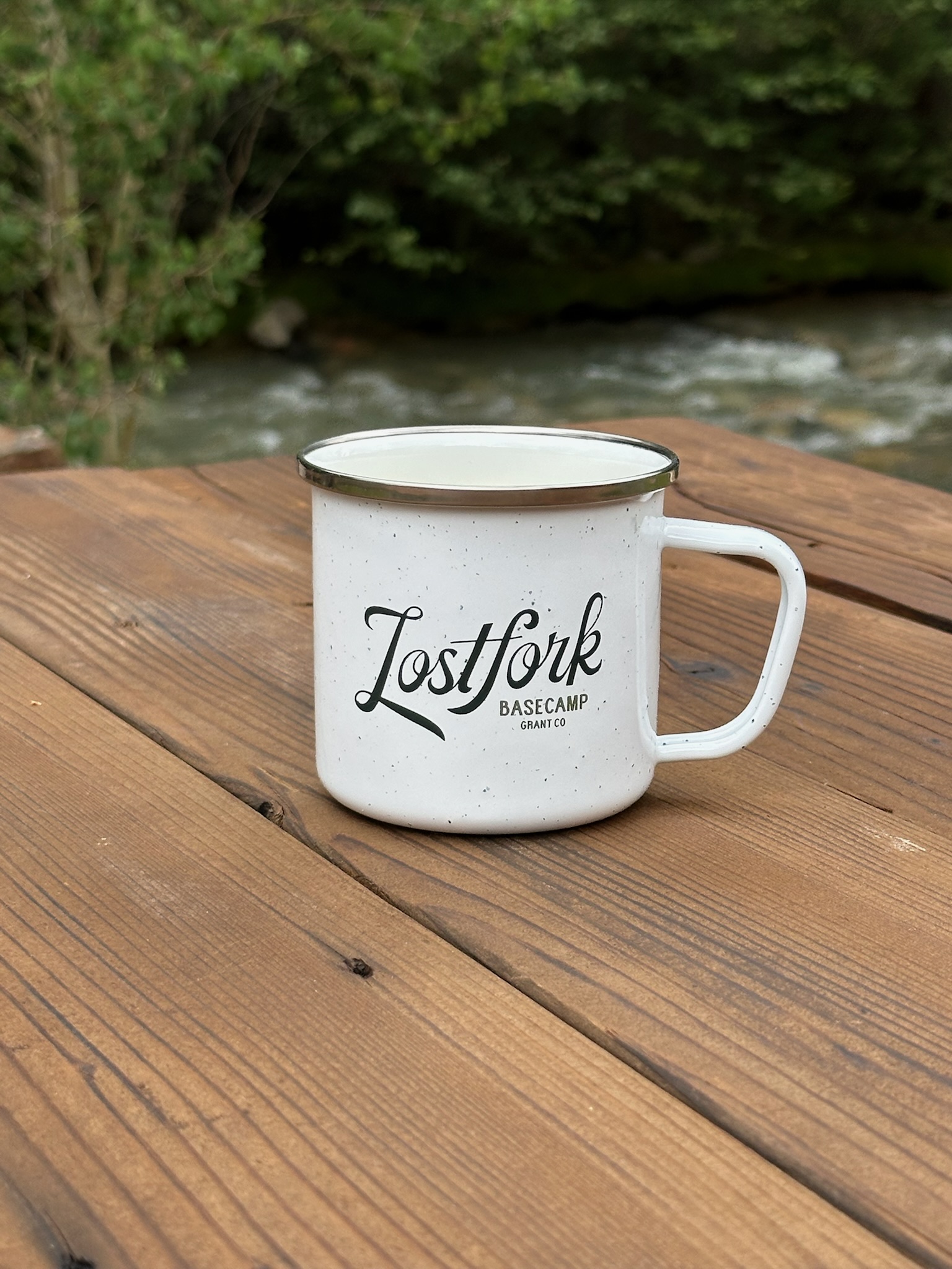 A white enamel mug with the words "Lost Fork Basecamp Grant Co." sits on a wooden table outdoors, with a river and green trees visible in the blurred background.