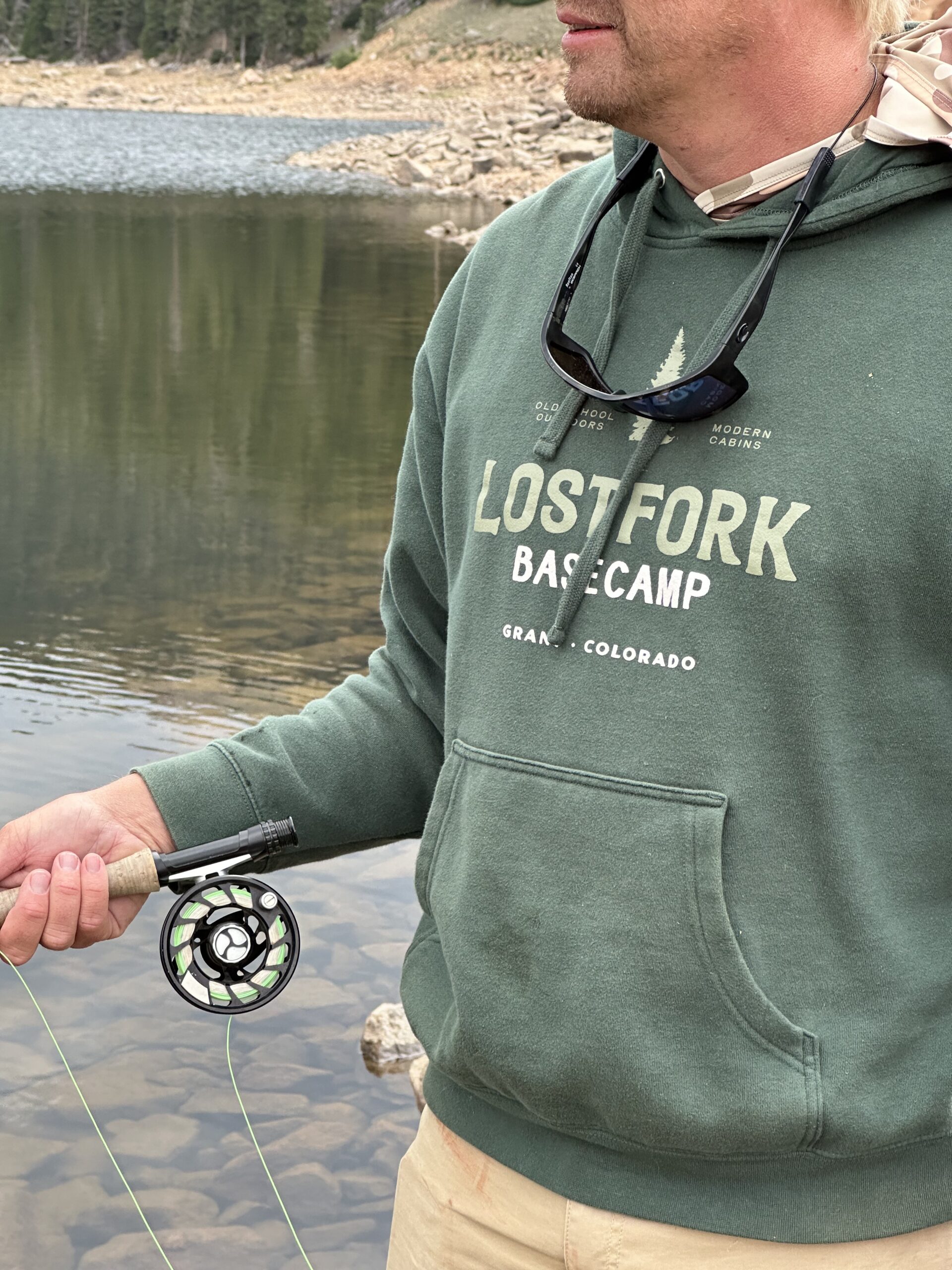 A person wearing a green "Lost Fork Basecamp" hoodie holds a fishing rod by a calm lakeshore, with rocks and trees in the background. Sunglasses hang from their neck.