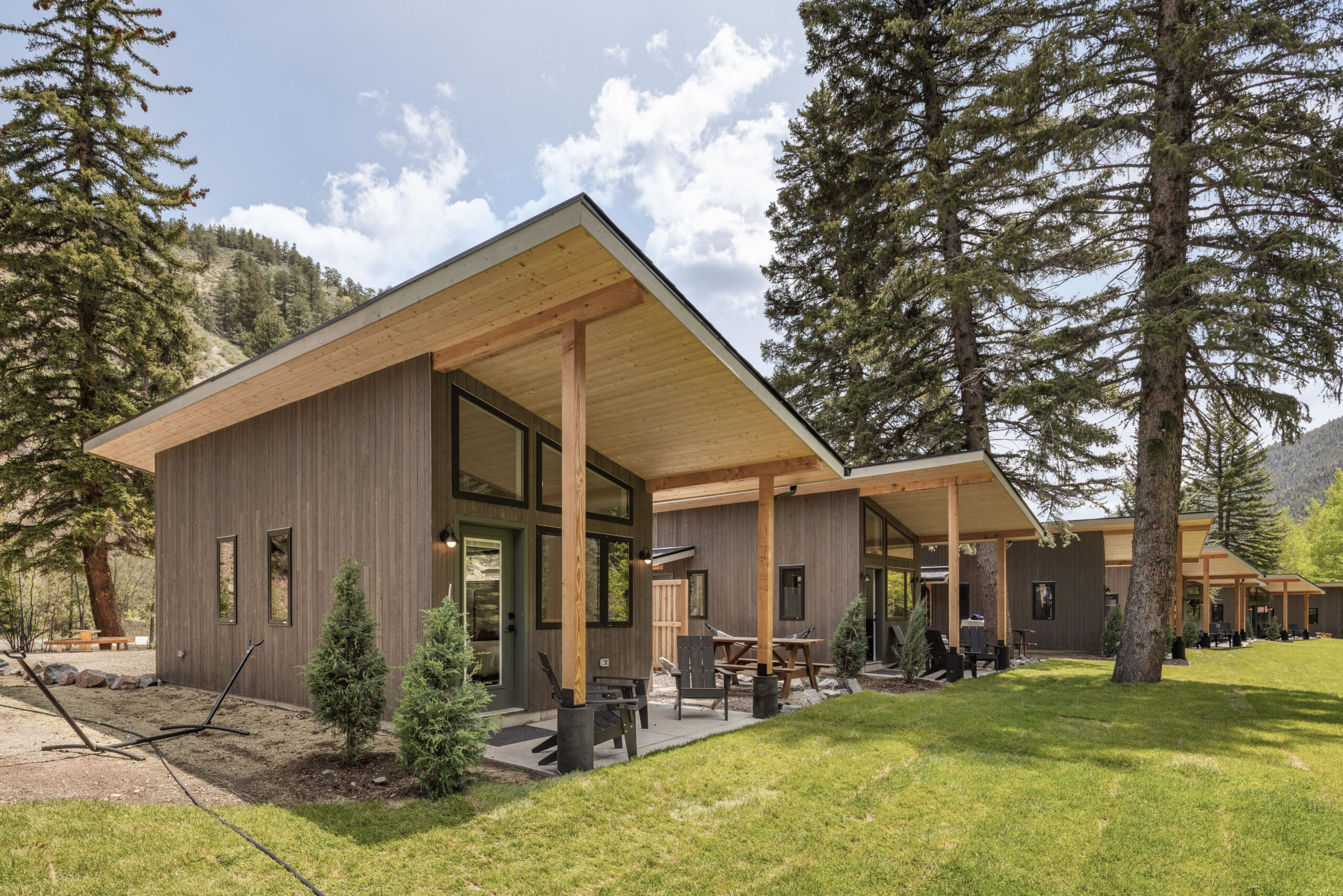 Modern wooden cabins with slanted roofs and large windows sit among tall pine trees, with patios featuring outdoor seating. Lush green grass covers the ground, and forested hills rise in the background under a partly cloudy sky.