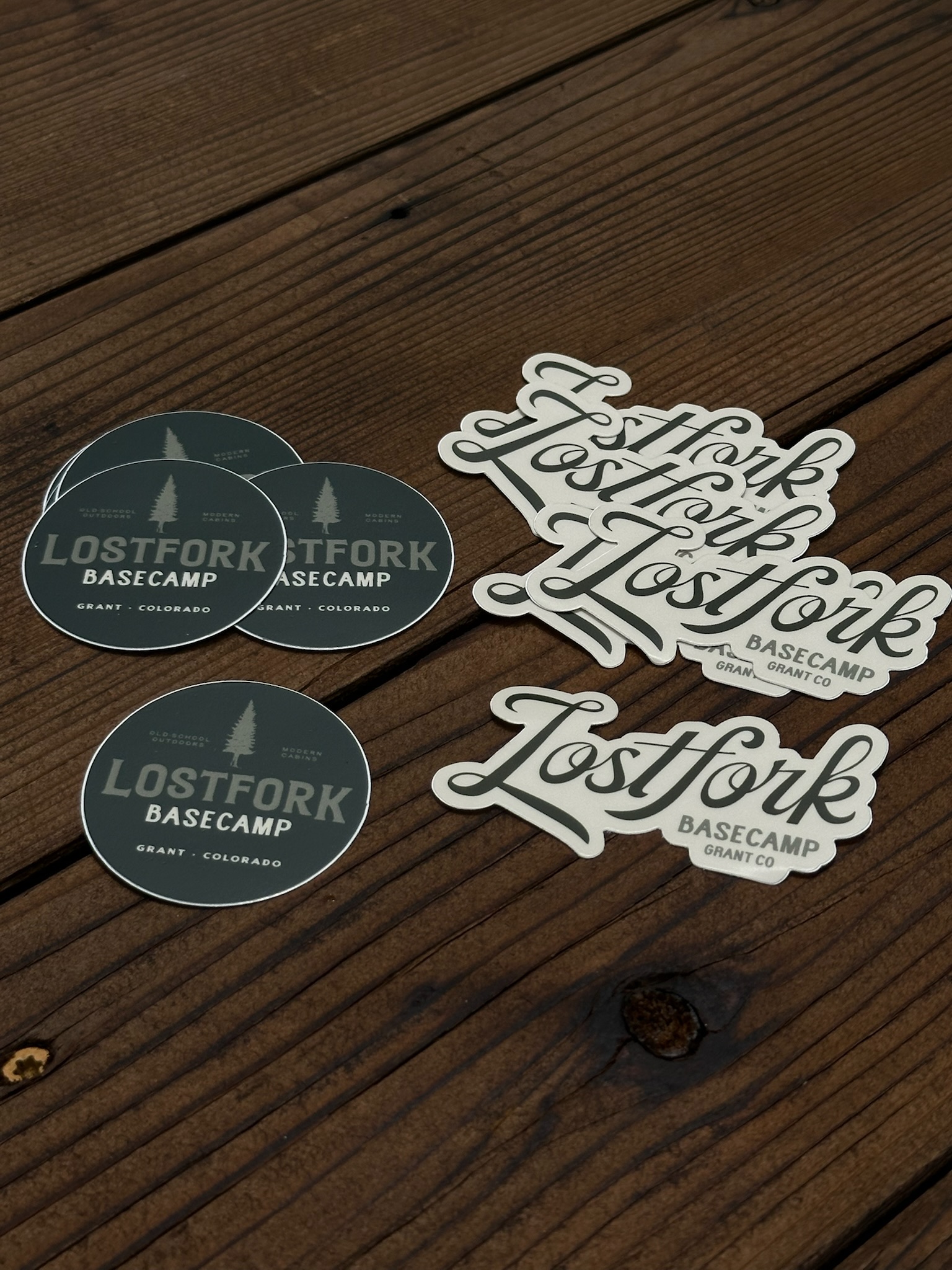 Stickers on a wooden surface: four round stickers reading “Lostfork Basecamp Grant, Colorado” and four white script stickers also reading “Lostfork Basecamp Grant, CO.”.