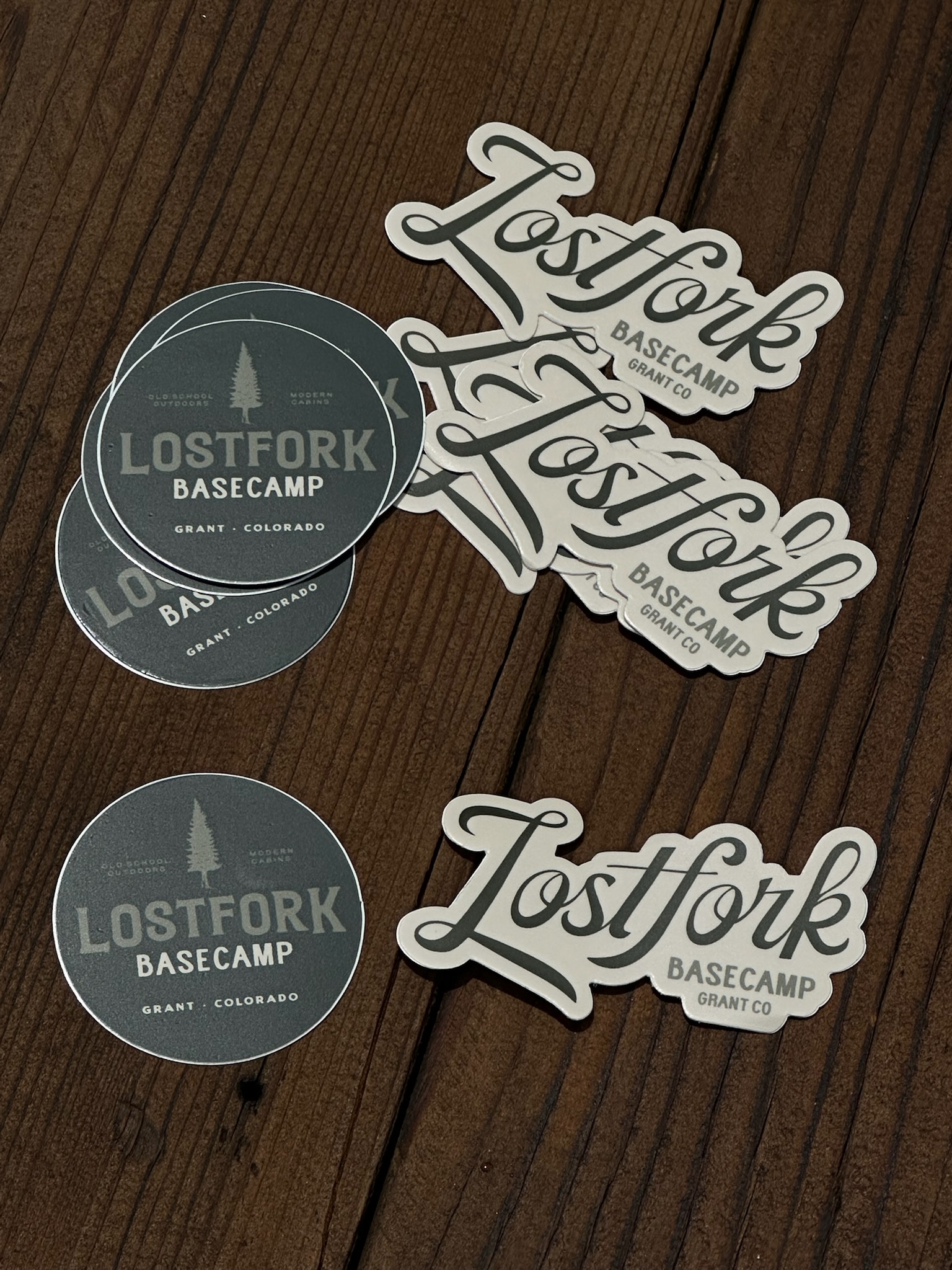 A group of circular and script-shaped stickers on a wooden surface. The stickers read "Lostfork Basecamp Grant, CO" in various fonts and include a small pine tree graphic.