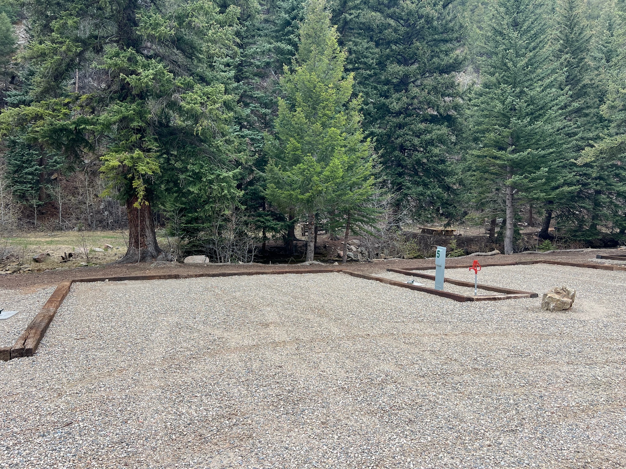 A gravel campsite with wooden borders is set among tall evergreen trees. A utility hook-up and red water spigot are visible, and a small stream flows in the background amid the forested landscape.