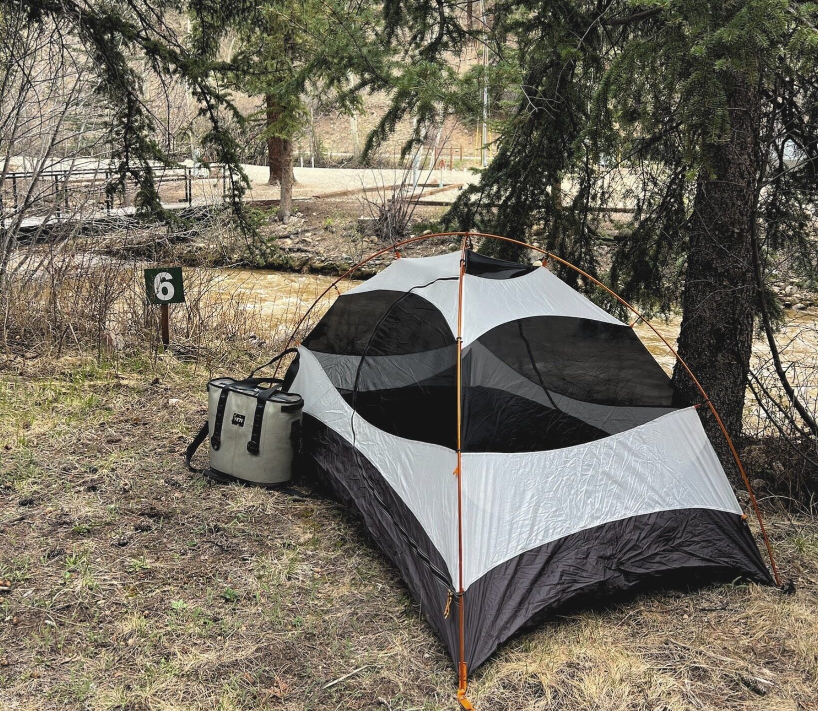 A gray and black tent is set up under a tree at campsite number 6, with a cooler next to it and a wooded area in the background.