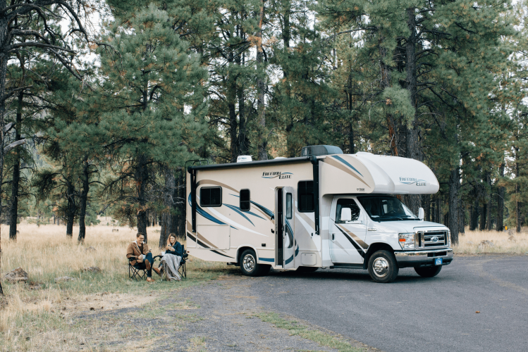 A large RV is parked near a forest with tall pine trees. Three people are sitting on camping chairs beside the RV, enjoying the outdoors on a clear day.