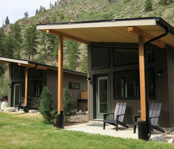 Two modern cabins with wooden frames and large windows sit on a grassy lawn with hills and trees in the background. Two black chairs are on the porch of each cabin.
