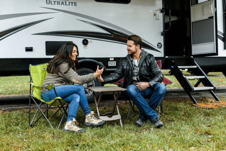 A smiling woman and man sit in camping chairs outside a camper van, holding hands across a small table, enjoying the outdoors on a grassy area.