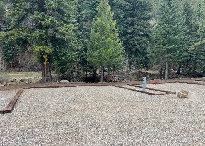 A gravel campsite with wooden borders is set among tall evergreen trees. A utility hook-up and red water spigot are visible, and a small stream flows in the background amid the forested landscape.