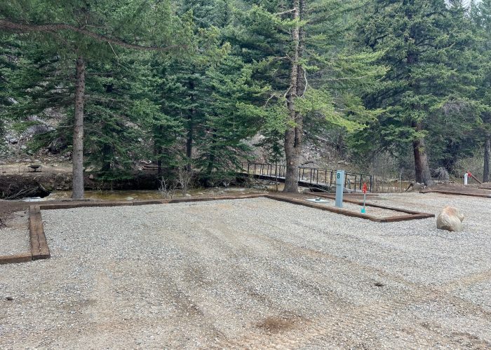 Gravel campsite with utility hookups surrounded by tall pine trees. A small wooden footbridge crosses a creek in the background, and a large rock sits near the edge of the site.
