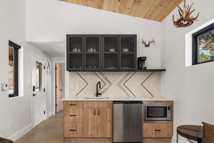 Modern kitchenette with wood cabinets, black fixtures, a geometric tile backsplash, glass-front upper cabinets, microwave, small refrigerator, antler decor, and natural light from windows.