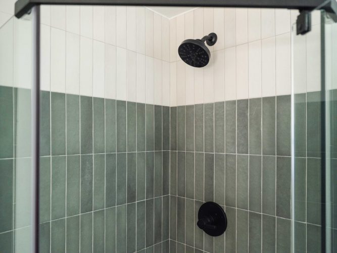 A shower with green and white vertical tile walls, featuring a black showerhead and matching black control knob. The shower is enclosed with glass panels.