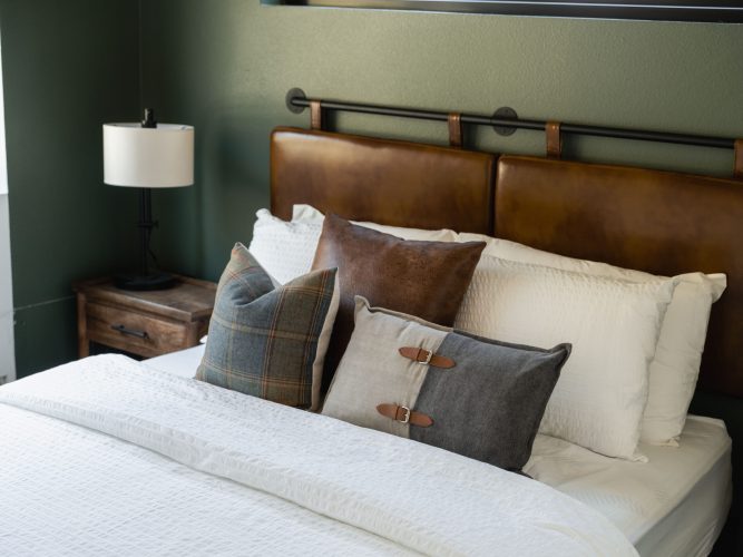 A neatly made bed with white bedding, three decorative pillows in plaid, brown, and gray, and a brown leather headboard; a wooden nightstand with a lamp sits beside the bed against a green wall.